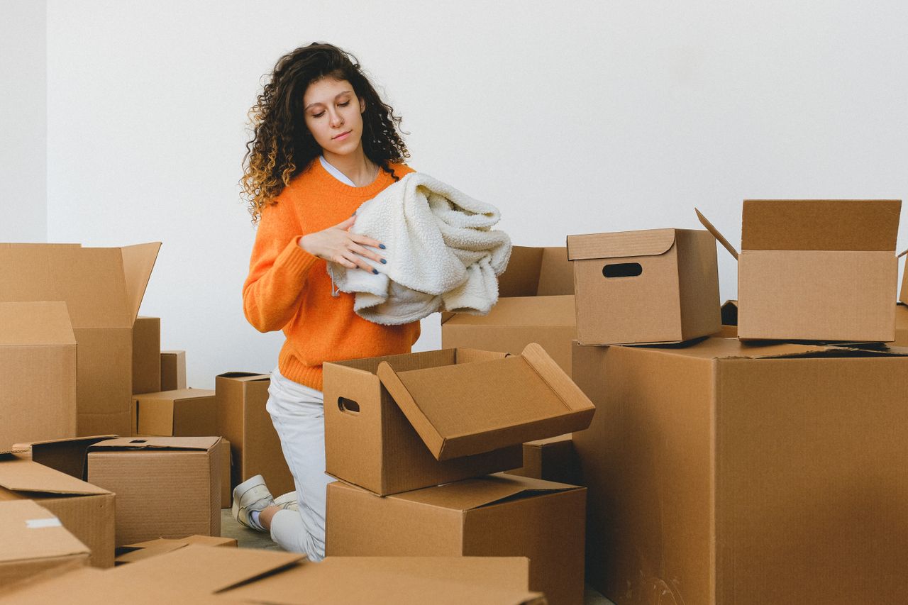 services-03 Concentrated young lady with curly hair standing on knees on floor and packing clothes into carton boxes before moving into new flat