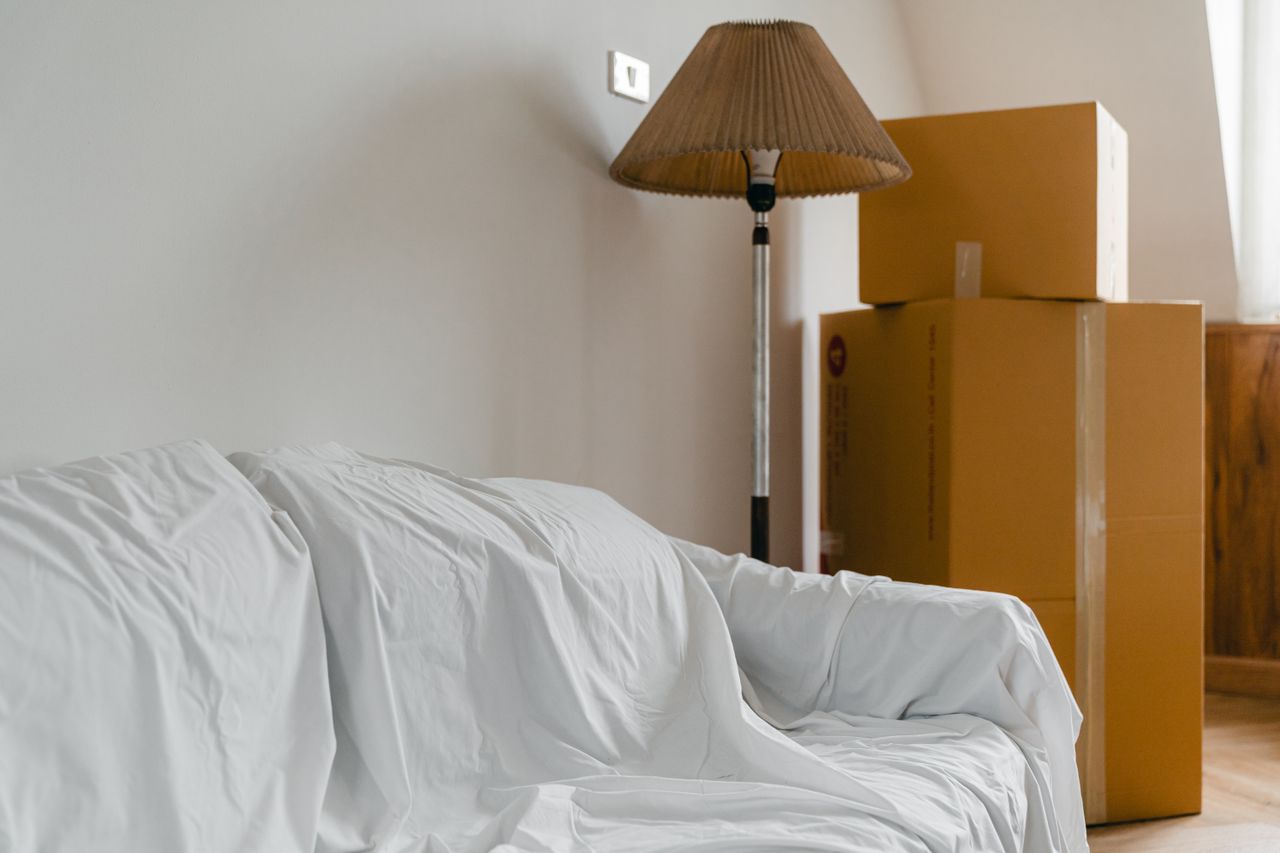 services-04 Covered with sheet sofa and floor lamp and packed cardboard boxes in light spacious room of new contemporary house